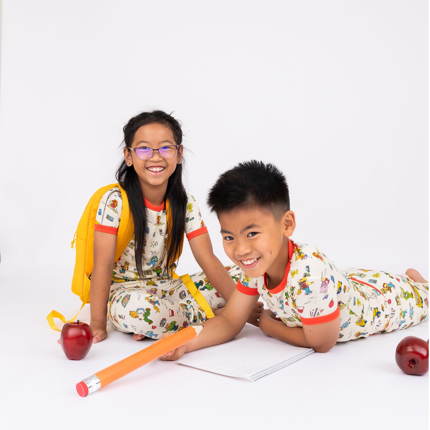 Sibling wearing our short Sleeve Kids Pajama Pants Set from buttery-soft bamboo, featuring red trims and playful illustrations of Arthur™ PBS Character, books, and school bus, a beige background.