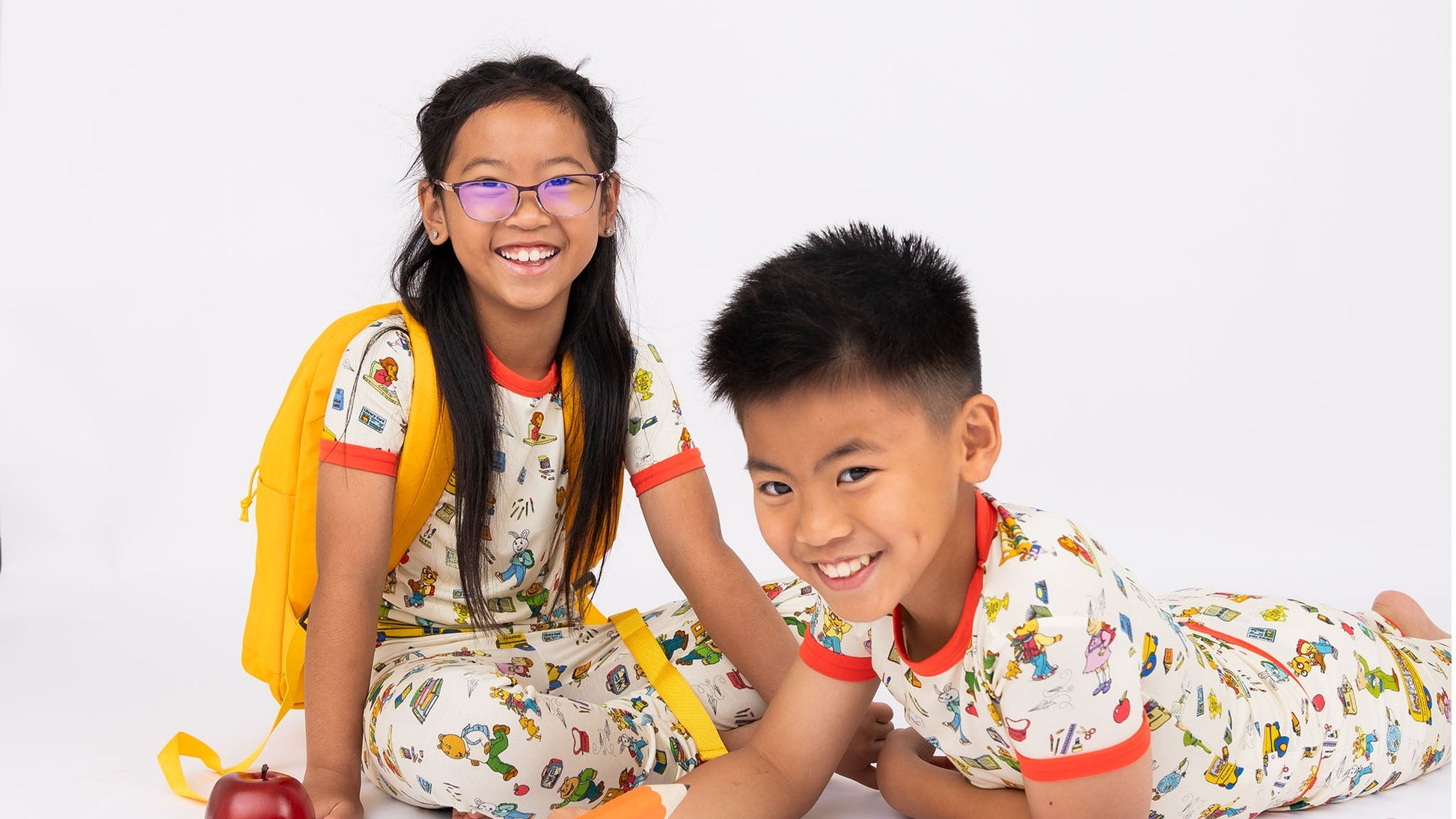 Sibling wearing our short Sleeve Kids Pajama Pants Set from buttery-soft bamboo, featuring red trims and playful illustrations of Arthur™ PBS Character, books, and school bus, a beige background.