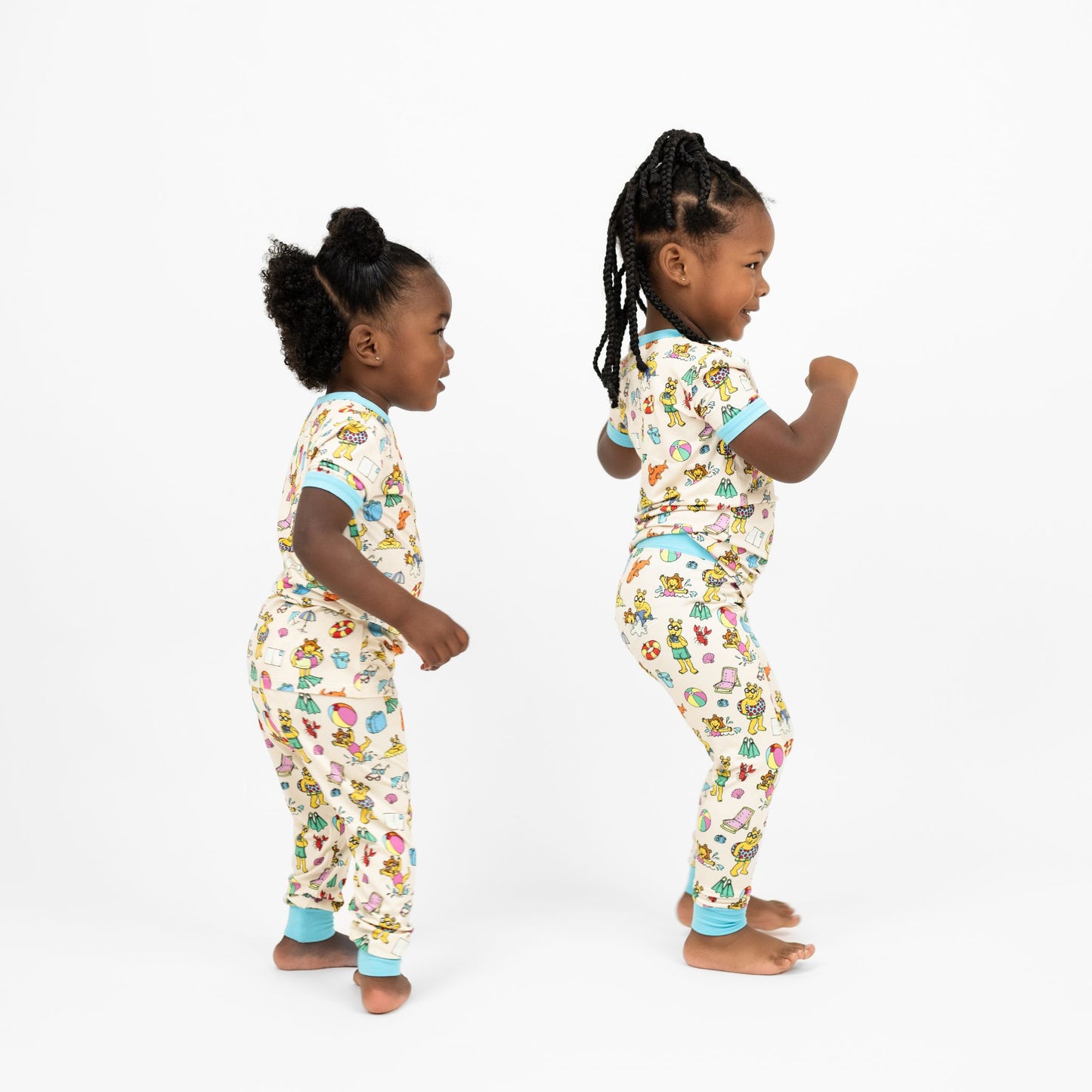 Matching siblings dancing and smiling wearing our Arthur's Family Vacation Bamboo Short Sleeve Kids Pajama Pants Set featuring vibrant light blue trim and a playful print of Arthur™ in swimwear surrounded by fun beach props — perfect for family getaways.
