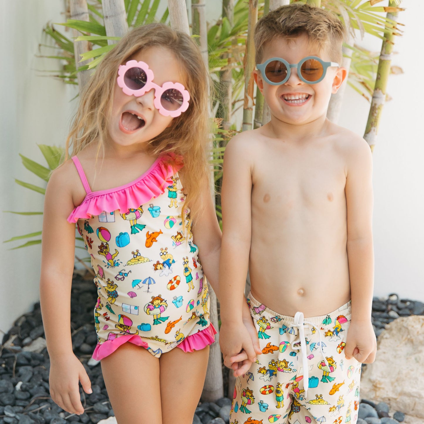 Siblings smiling with our Arthur's Family Vacation one piece and swim trunks with a playful print of Arthur™ in swimwear surrounded by fun beach props — perfect for family getaways.