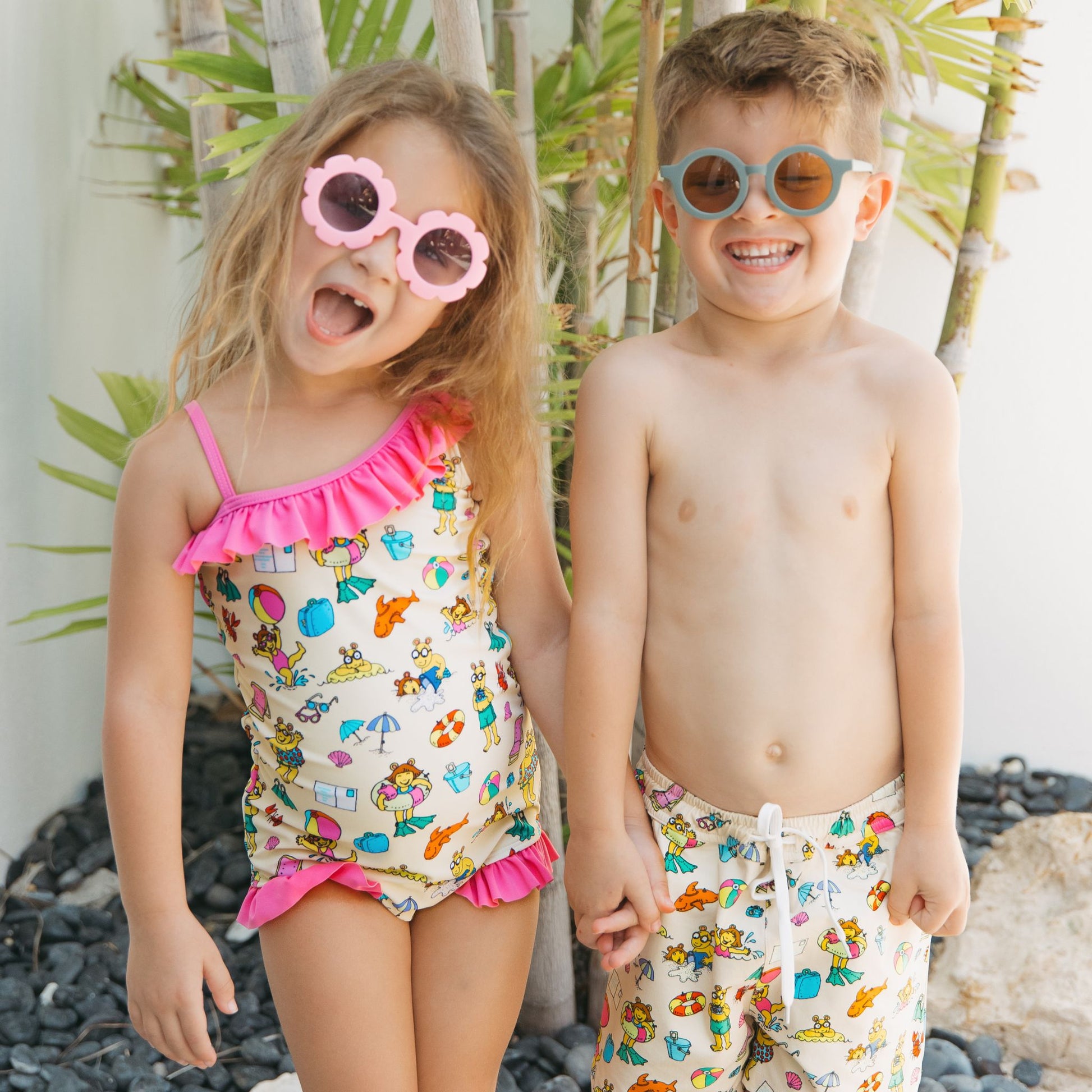 Siblings smiling with our Arthur's Family Vacation one piece and swim trunks with a playful print of Arthur™ in swimwear surrounded by fun beach props — perfect for family getaways.