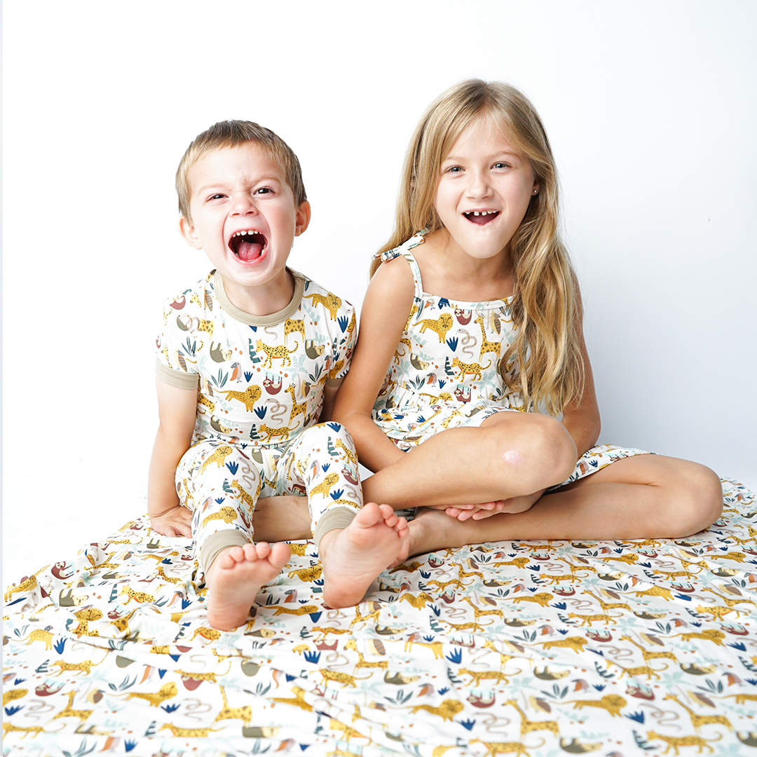 two kids sit on the "jungle friends" bamboo blanket. the boy on the left roars like a lion in his "jungle friends" 2-piece matching pajama set. the girl next to him laughs in her "jungle friends" dress. the "jungle friends" print is a mix of jungle animals that include giraffes, lions, cheetahs, sloths, snakes, and forest plants.