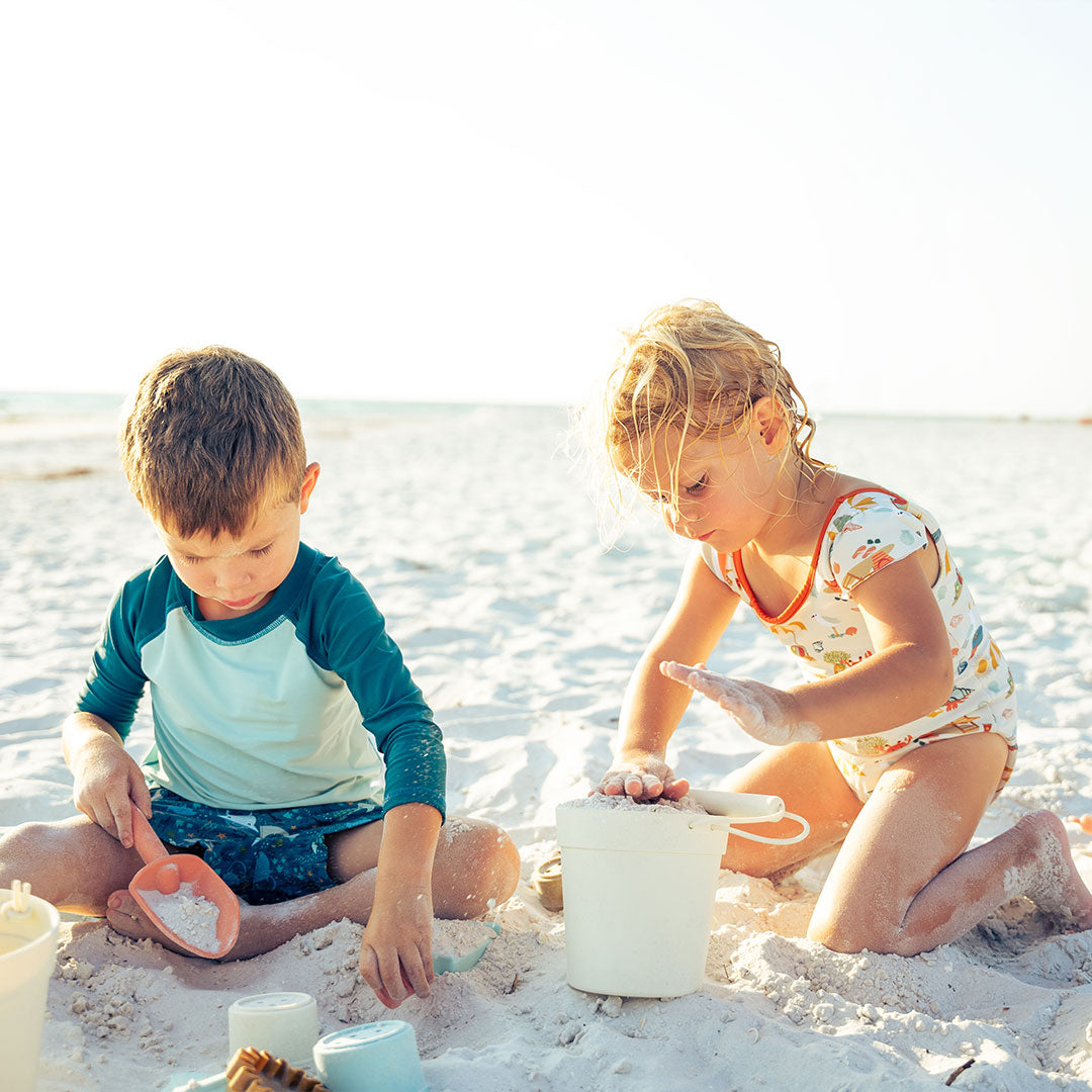 2 toddlers play in the sand with the silicone beach bucket toy play set.