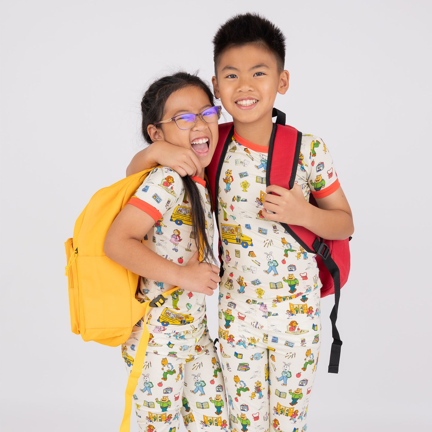 Siblings wearing the short Sleeve Kids Pajama Pants Set from buttery-soft bamboo, featuring red trims and playful illustrations of Arthur™ PBS Character, books, and school bus, a beige background.