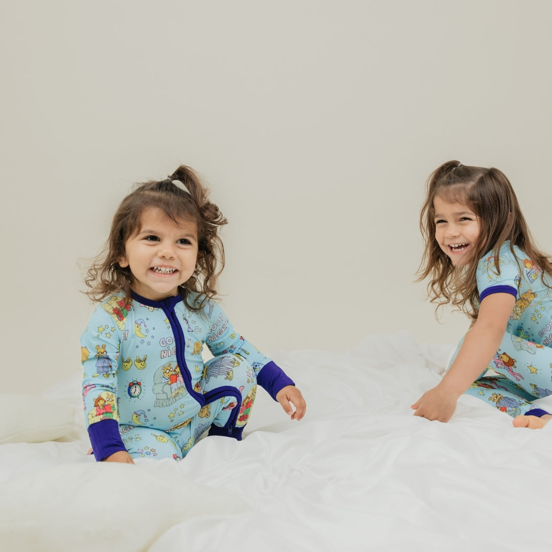 Adorable moment with sisters smiling. She is wearing our collaboration with PBS Good Night Arthur™ pattern. The pajama features Arthur and friends in a cozy bedtime scene with storybooks, toothbrushes, bunny slippers, a sleeping puppy, crescent moons wearing sleep caps, twinkling stars, and the words "GOOD NIGHT!" on a soft blue background.