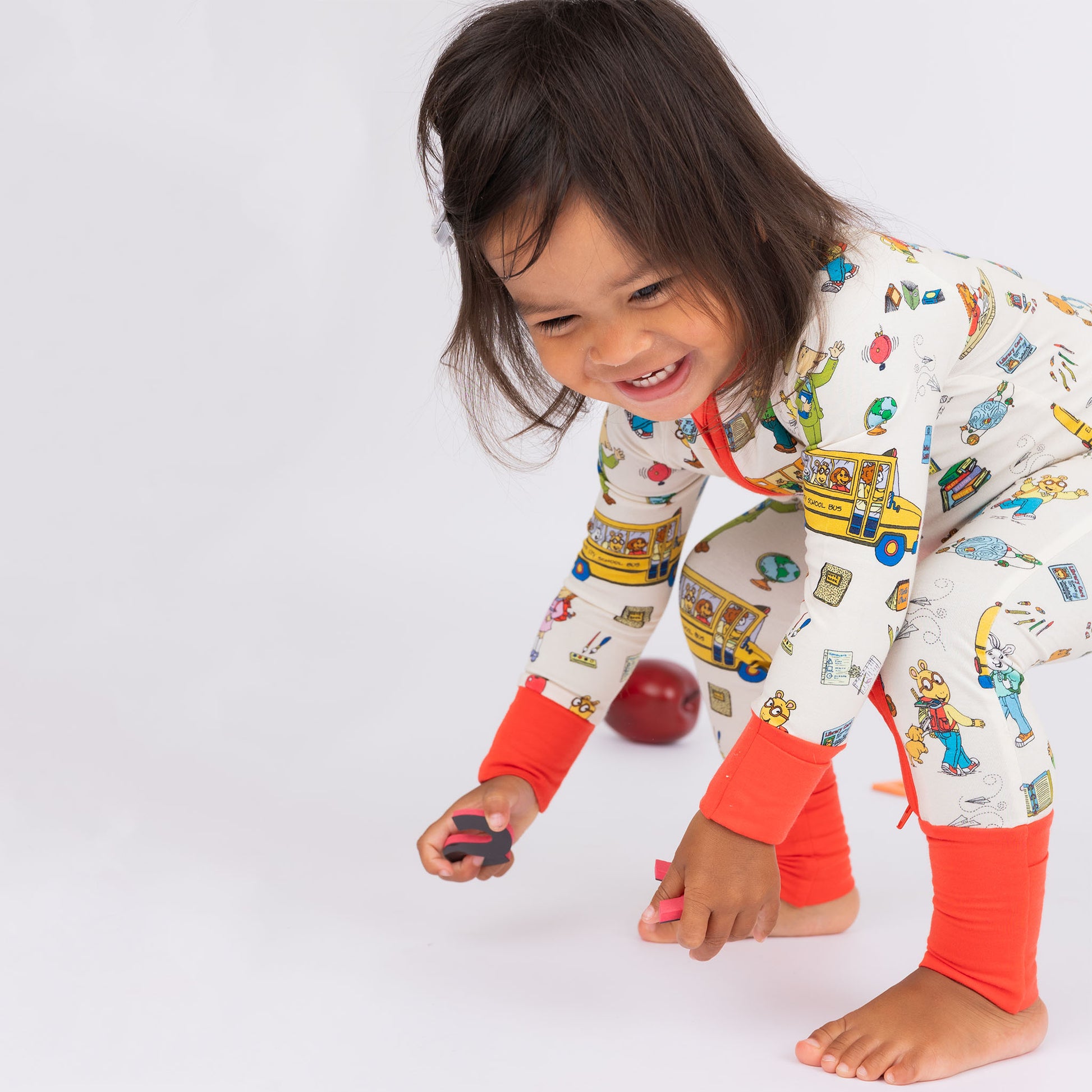 Baby girl playing while wearing our buttery-soft  bamboo convertible pajama featuring red trims and playful illustrations of Arthur™ PBS Character, books, and school bus, a beige background.