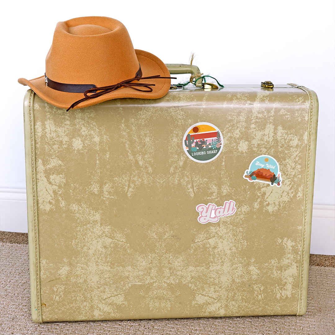 an old-fashion suitcase with a cowboy hat placed on top of it. there are 3 stickers, one that says "y'all" one that says "stay wild," and one that says "looking sharp"