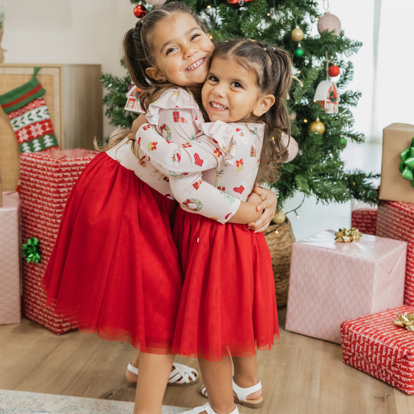 Sisters hugging by the Christmas tree in matching Sugar Plum bamboo tulle dresses, perfect for festive celebrations.