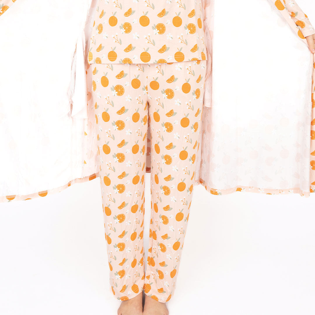 someone posing in the "freshly squeezed" bamboo lounge relaxed pants. they are also wearing the freshly squeezed women's top and spreading open their "freshly squeezed" robe to show her clothes underneath. the "freshly squeezed" print has an assortment of full and half cut oranges scattered around. there is also flower heads and flower stems that intermingle within the print. this is all space out around a pink background space.