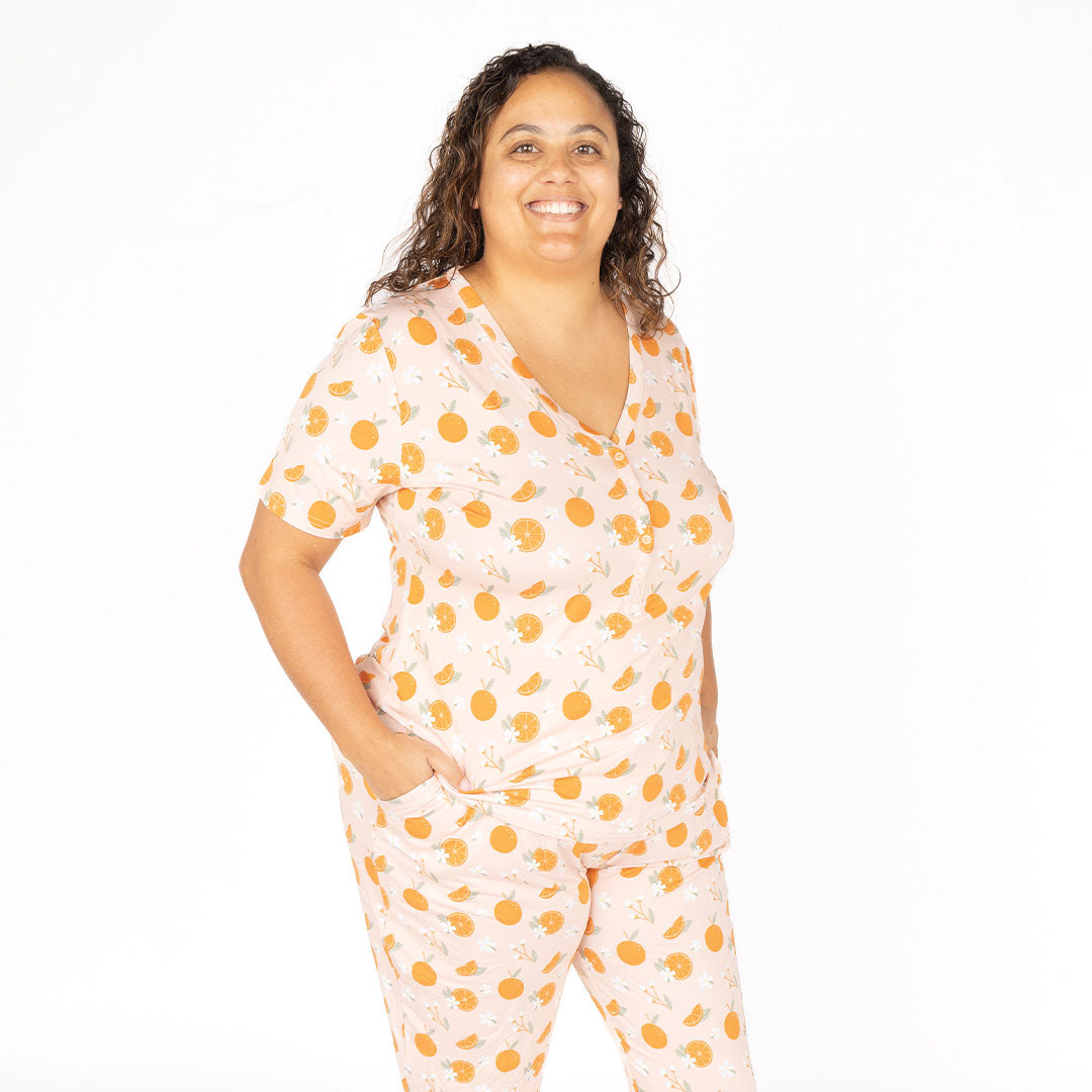 a woman posing in her "freshly squeezed" women's top. the "freshly squeezed" print has an assortment of full and half cut oranges scattered around. there is also flower heads and flower stems that intermingle within the print. this is all space out around a pink background space.