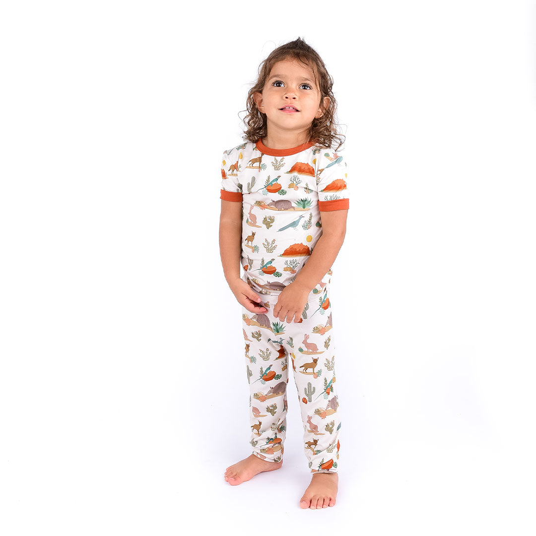 toddler girl stands wearing the "desert friends" 2-piece matching pajama set. the "desert friends" print is a scattered pattern of armadillo, cacti, other desert plants, rocks, desert foxes, lizards, and desert birds. this is all on a beige background.