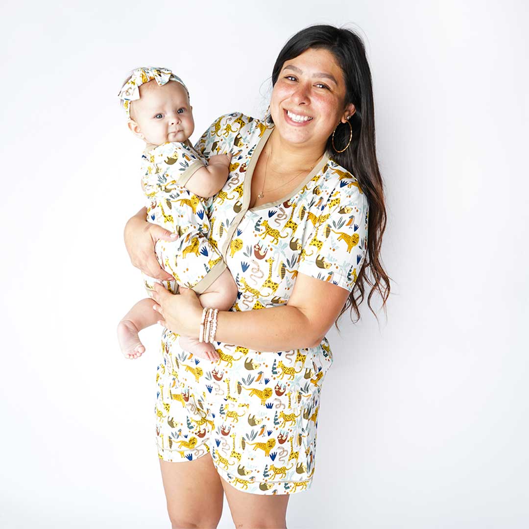 mom smiles and holds her baby. they are wearing matching "jungle friends" prints. the mom wears the women's shorts and top. the baby wears the shortie romper and headband. the "jungle friends" print is a mix of jungle animals that include giraffes, lions, cheetahs, sloths, snakes, and forest plants.