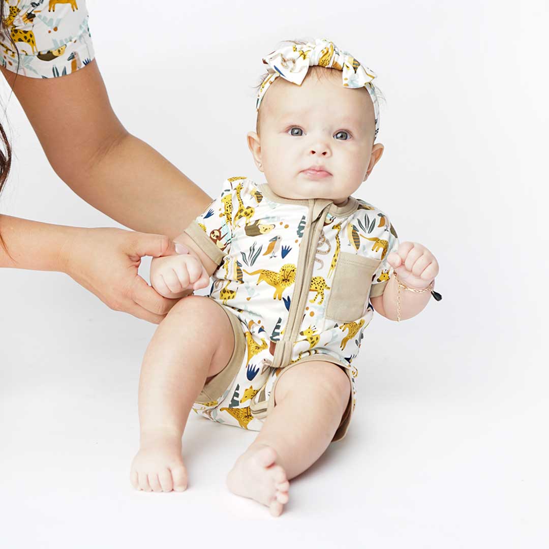 arms hold up a baby who is wearing the "jungle friends" shortie and headband. the "jungle friends" print is a mix of jungle animals that include giraffes, lions, cheetahs, sloths, snakes, and forest plants.