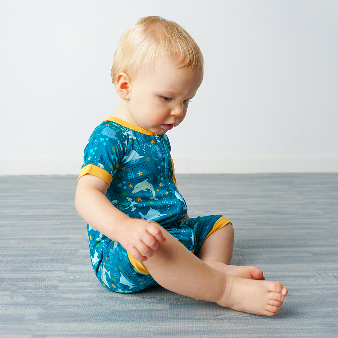 baby sits on the floor in his "ocean friends" bamboo baby boy shortie. the "ocean friends" print is a combination of dolphins, stingrays, fish, starfish, coral, bubbles, and sharks, all spread out on a deep sea blue background.