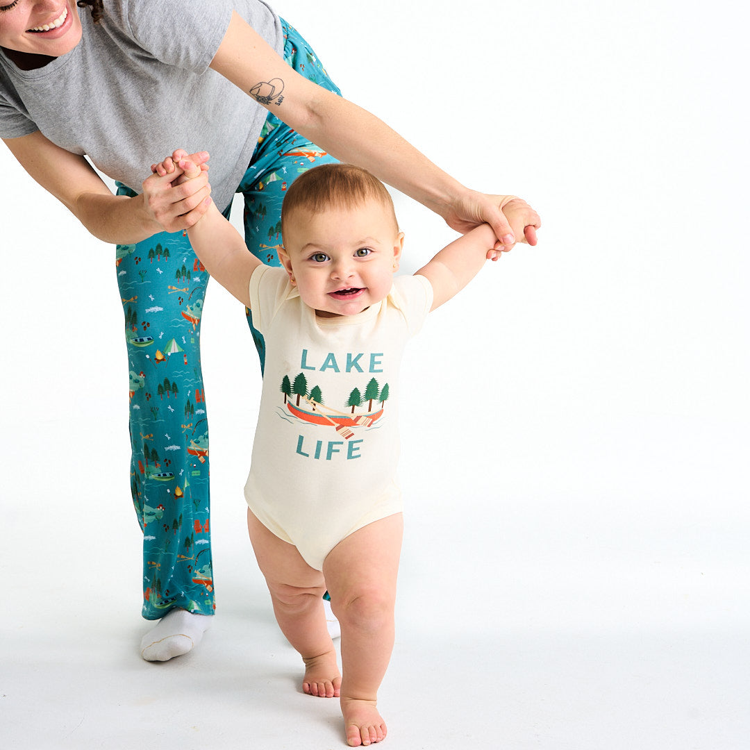 mom holds her baby up by their arms. the baby is in the "lake life" cotton onesie. this design says the phrase "lake life" and has a canoe, ores, oak trees, and some lake waves.