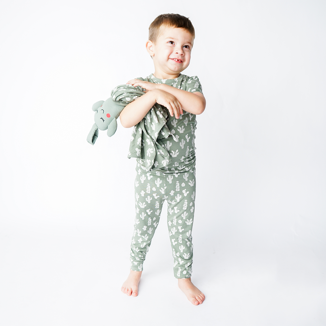 a young boy in the "stay sharp" matching pajama set, holds the "stay sharp" lovey. the "stay sharp" print is a variety of different white cacti on a greyish/green background.