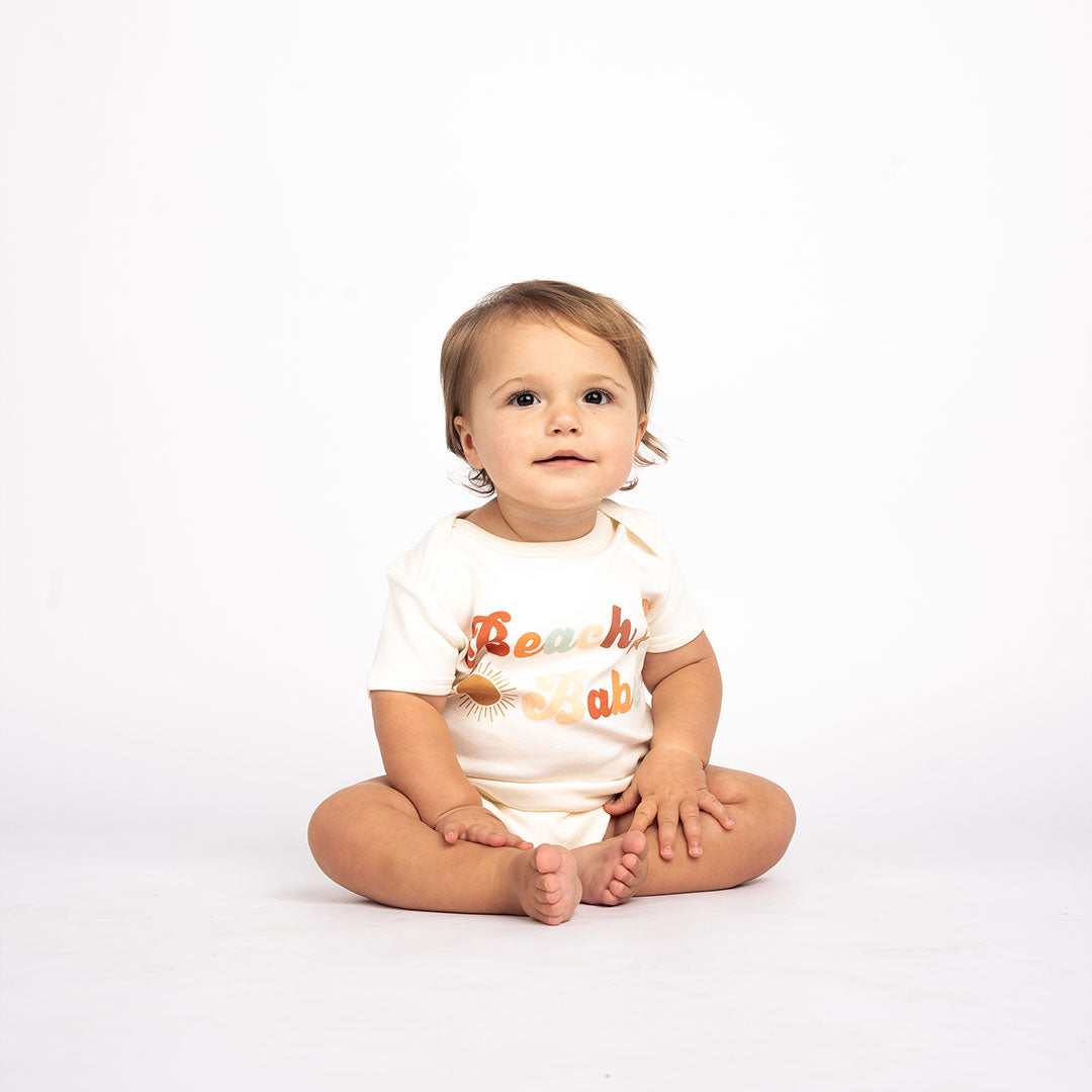 a baby sitting on the floor in the "beach babe" cotton onesie. the word 'beach babe' is spelt out in a groovy font with a sun, starfish, and shell surrounding it.