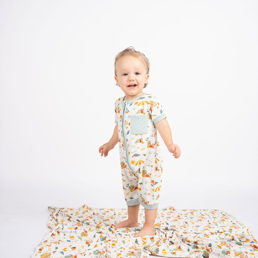 a baby in the "beach days" shortie. he is standing on the "beach days" blanket. the "beach days" print is a scattered print of beach umbrellas, beach balls, sand buckets and shovels, coral, shells, beach chairs, seagulls, flipflops, and sunglass mixed in a orderly and creative way.
