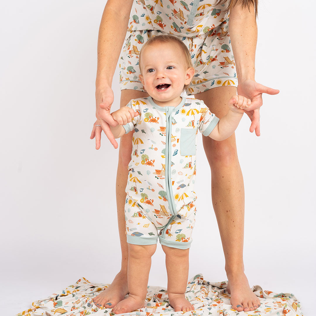 a mom standing, bent over, and holding her baby up by his hands. the baby is smiling. they are all wearing "beach days" apparel. the baby is in the shortie and the mom is in the women's shorts and the women's top. the "beach days" print is a scattered print of beach umbrellas, beach balls, sand buckets and shovels, coral, shells, beach chairs, seagulls, flipflops, and sunglass mixed in a orderly and creative way.