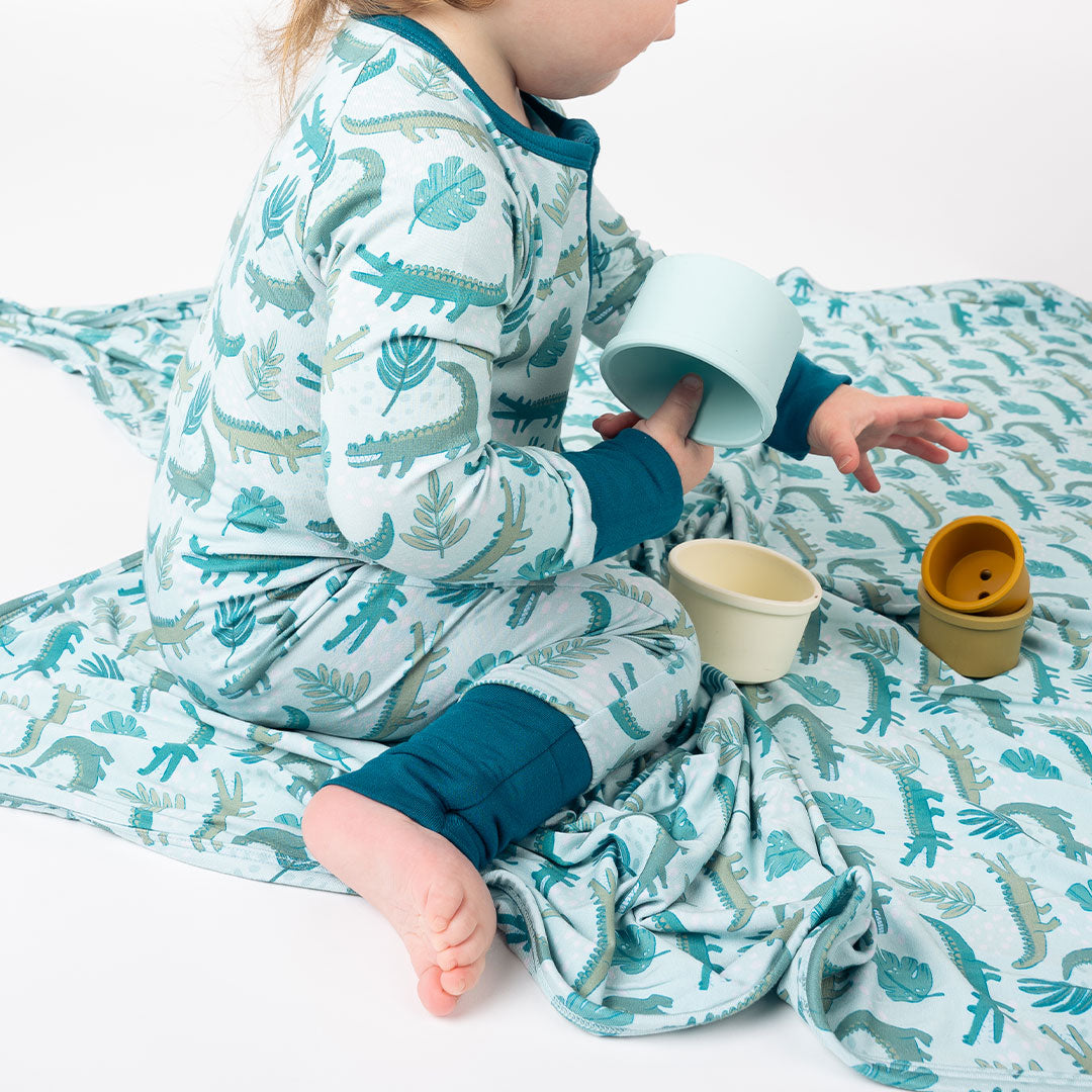 up close photo of a toddler playing with the silicone cups, sitting on the "later gator" blanket. the "later gator" print has a mix of light and dark green alligators, leaves, and white dots scattered on a teal colored background.