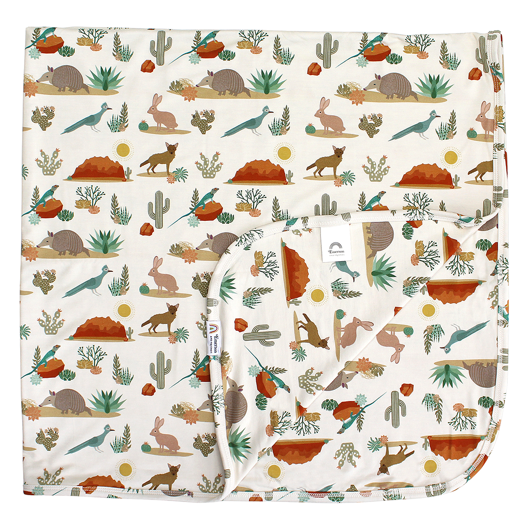 the "desert friends" bamboo blanket. the "desert friends" print is a scattered pattern of armadillo, cacti, other desert plants, rocks, desert foxes, lizards, and desert birds. this is all on a beige background.