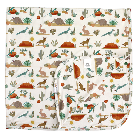 the "desert friends" bamboo blanket. the "desert friends" print is a scattered pattern of armadillo, cacti, other desert plants, rocks, desert foxes, lizards, and desert birds. this is all on a beige background.
