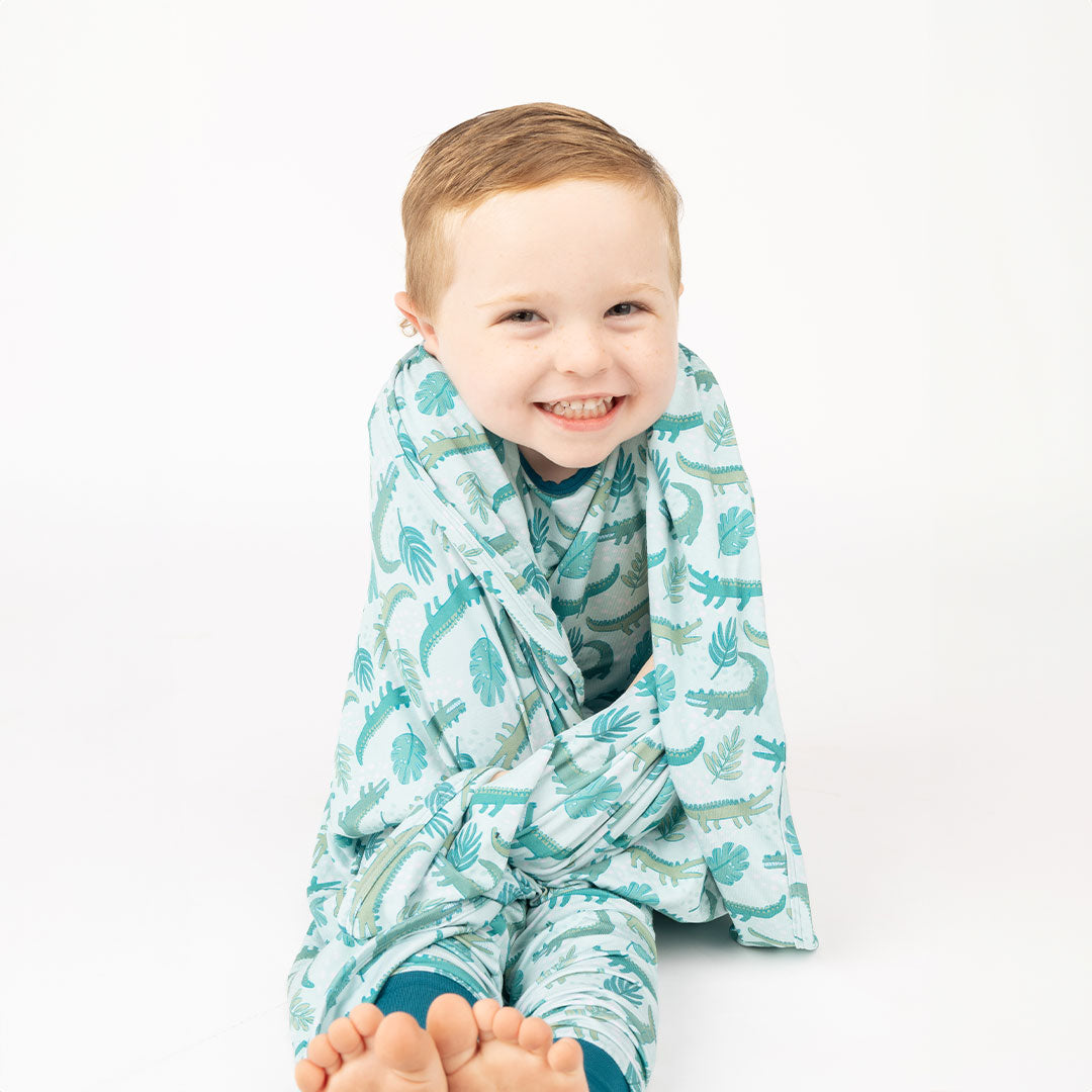 little toddler boy smiling big in his "later gator" convertible and his "later gator" blanket. the "later gator" print has a mix of light and dark green alligators, leaves, and white dots scattered on a teal colored background.