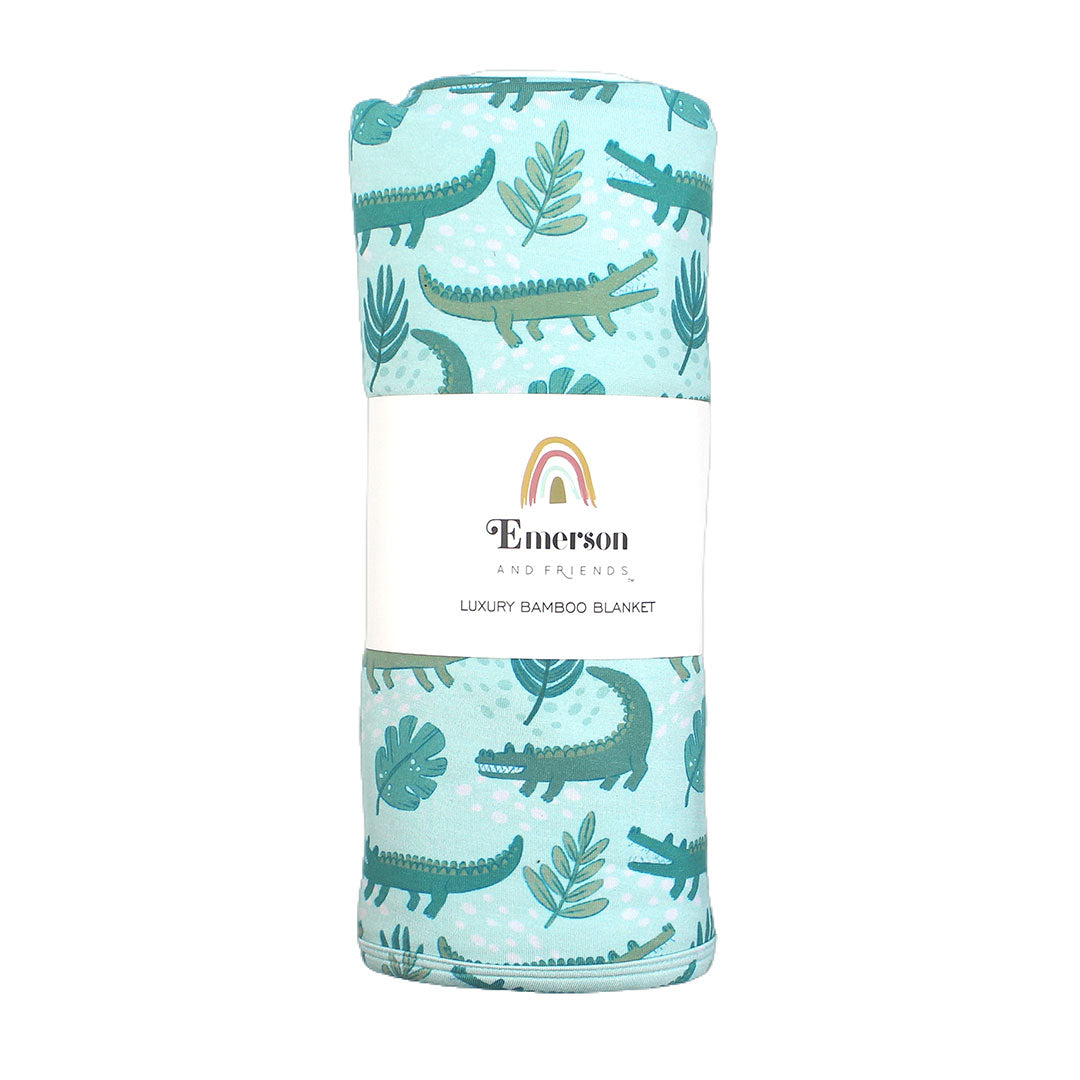 the "later gator" blanket in it's wrapped up packaging. the "later gator" print has a mix of light and dark green alligators, leaves, and white dots scattered on a teal colored background.