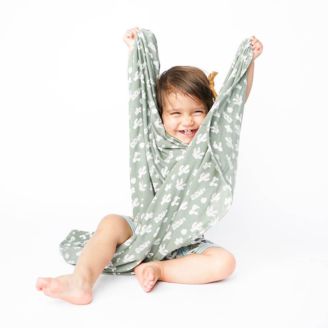 little baby sits on the floor and covers themself with the "stay sharp" bamboo blanket. the "stay sharp" print is a variety of different white cacti on a greyish/green background.