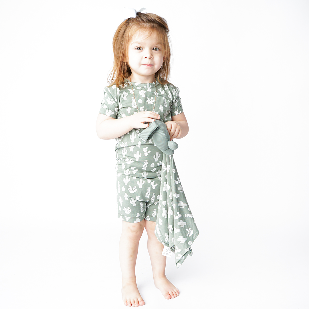 girl stands and holds the "stay sharp" lovey. she is wearing the "stay sharp" matching pajama set. the "stay sharp" print is a variety of different white cacti on a greyish/green background.