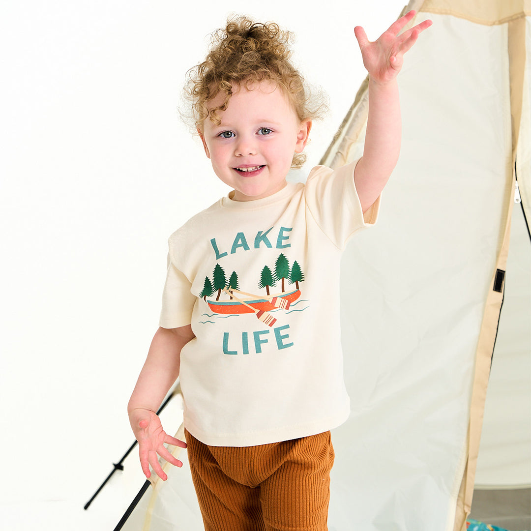 young child wears the "lake life" cotton tee. this design says the phrase "lake life" and has a canoe, ores, oak trees, and some lake waves.