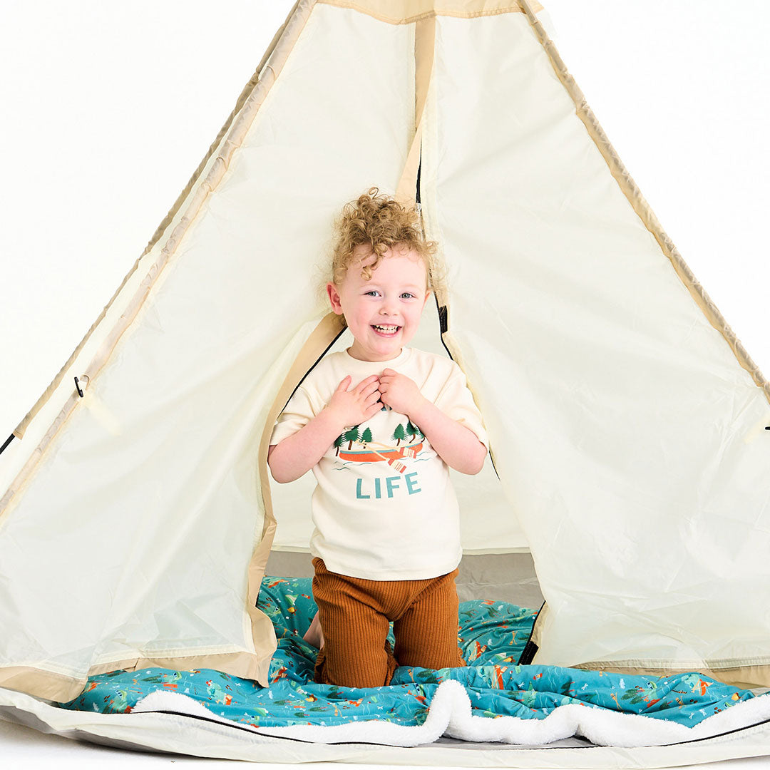 little kid sits in a tent. they wear the "lake life" tee. this design says the phrase "lake life" and has a canoe, ores, oak trees, and some lake waves.