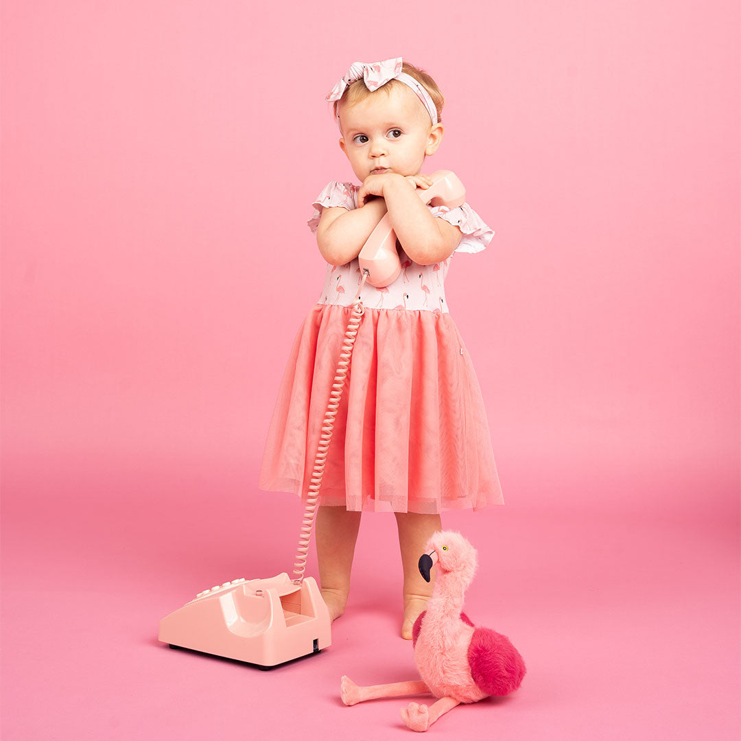 little toddler girl in her "fancy flamingos" twirl dress. she is holding an old-school phone and "Florence the flamingo" is sitting next to her. the "fancy flamingos" print is a pattern of multiple pink flamingoes scattered around the print.