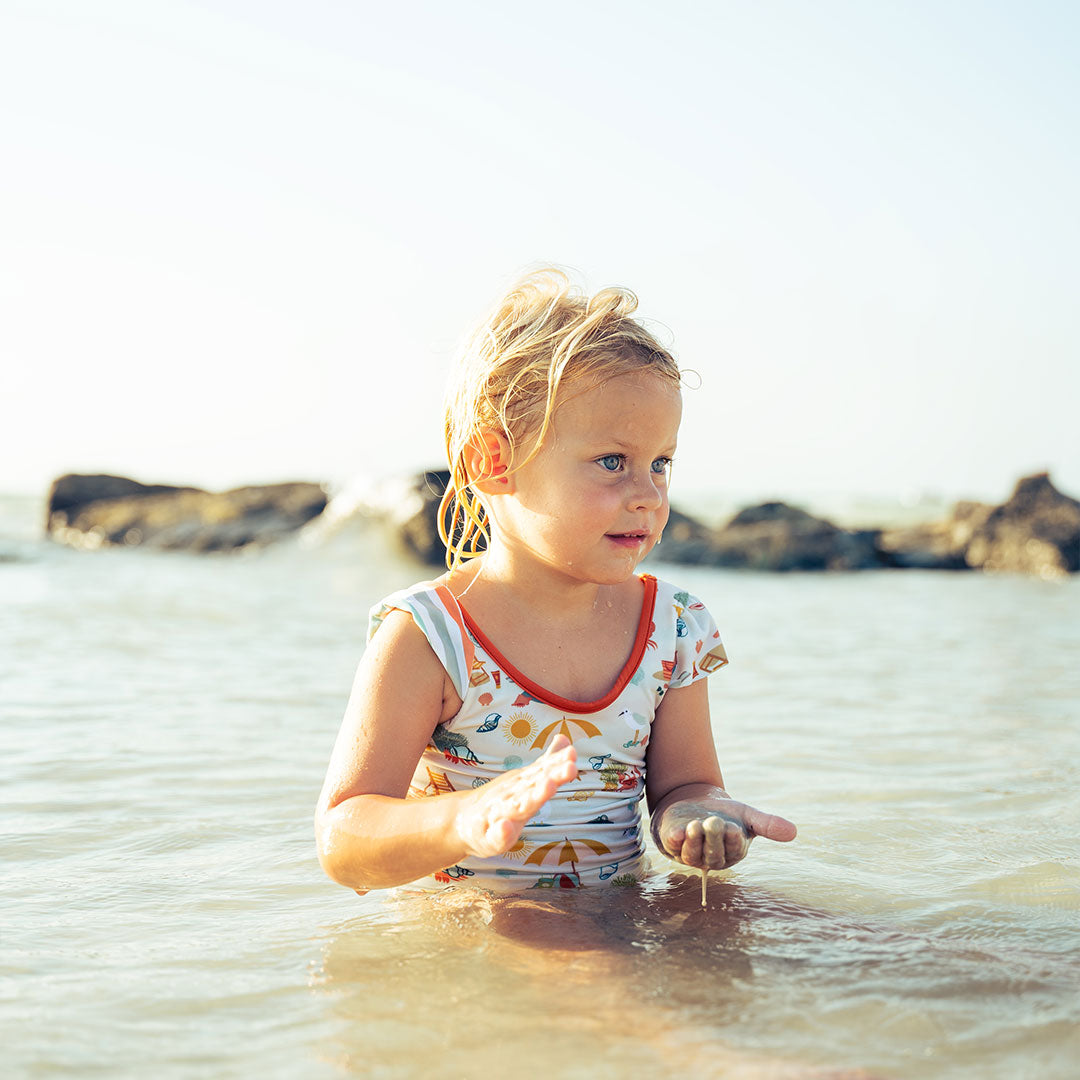 girl sits in the water, playing with sand in her hands. she is wearing the "beach days" reversible one piece ruffled sleeved swim suit. the "beach days" print is a scattered print of beach umbrellas, beach balls, sand buckets and shovels, coral, shells, beach chairs, seagulls, flipflops, and sunglass mixed in a orderly and creative way.