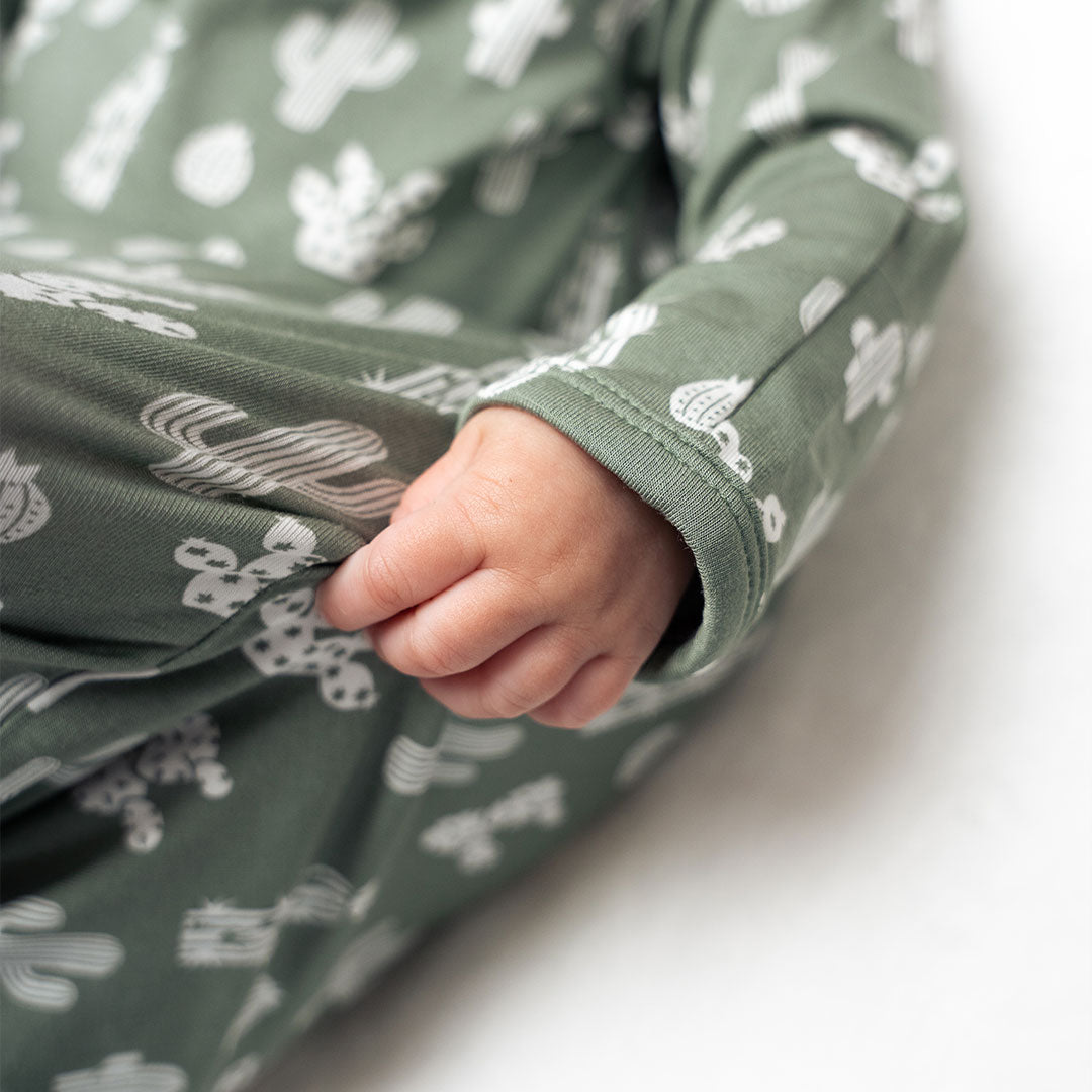 up close and zoomed in photo of a baby's hand. the baby is wearing the "stay sharp" gown. the "stay sharp" print is a variety of different white cacti on a greyish/green background.
