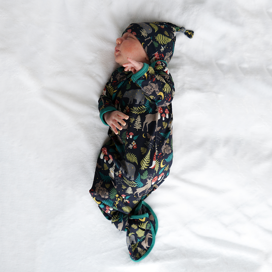 a infant baby is seen laying on his side on a white blanket. the baby is in the "night forest" gown and hat set. the "night forest" print is a night time themed design. you can see an array of forest animals ranging from, deer, bears, bunnies, birds, forest trees and leaves, flowers, and mushrooms. there are also starts and moons scattered around to enhance the night time atmosphere.