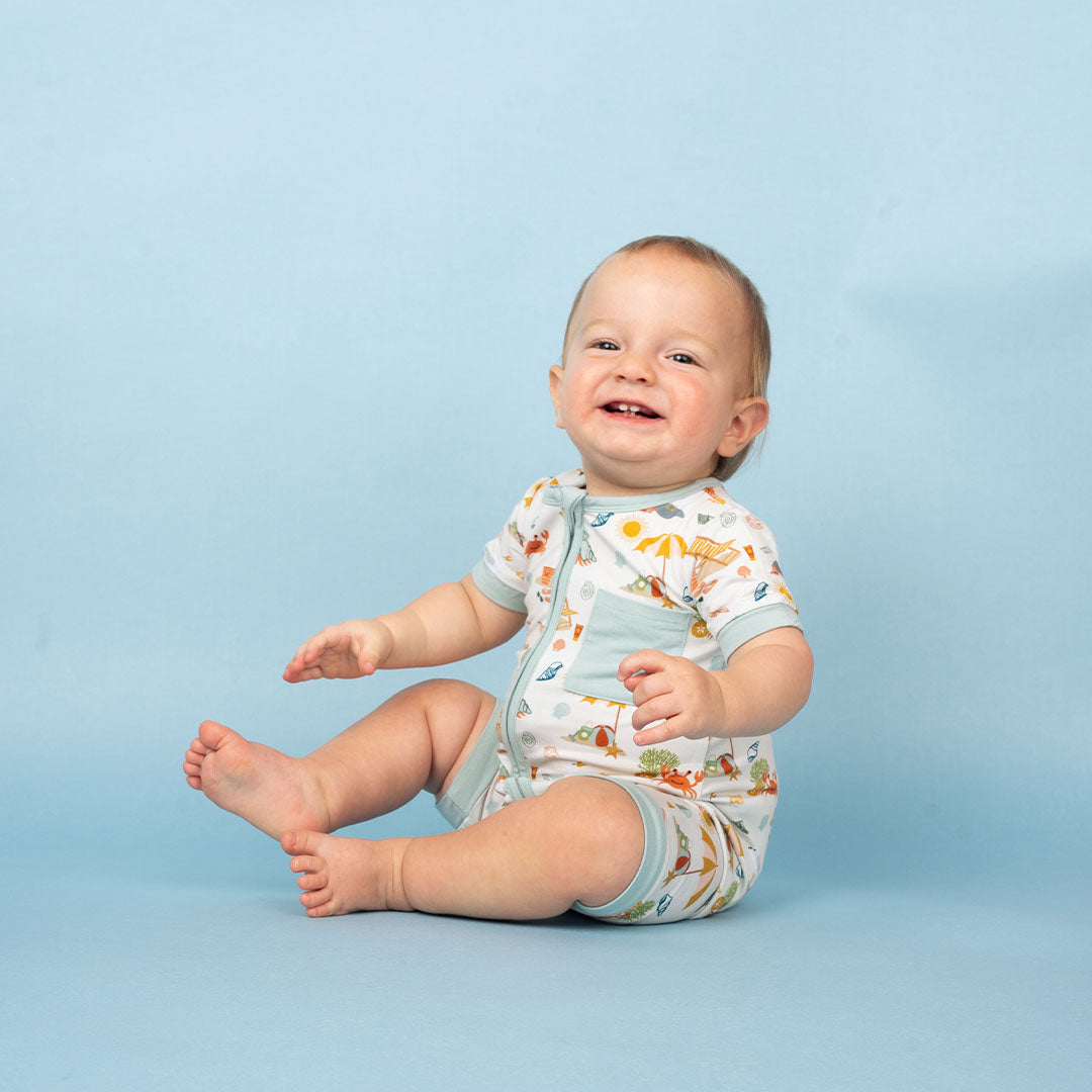 baby sits on the floor laughing in the "beach days"shortie romper. the "beach days" print is a scattered print of beach umbrellas, beach balls, sand buckets and shovels, coral, shells, beach chairs, seagulls, flipflops, and sunglass mixed in a orderly and creative way.