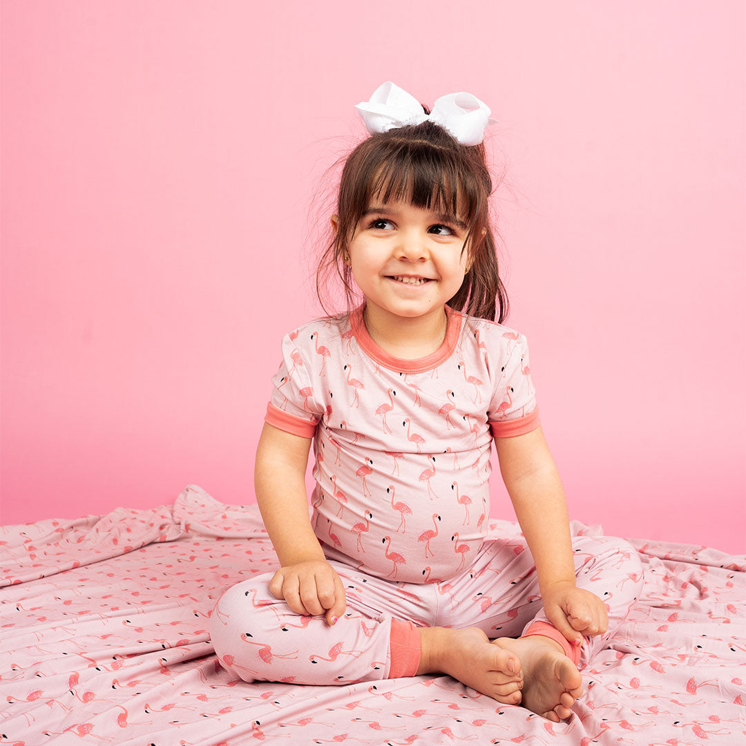 a cute toddler sitting on the "fancy flamingos" blanket, while wearing her "fancy flamingos" 2-piece matching pajama set. the "fancy flamingos" print is a pattern of multiple pink flamingoes scattered around the print.