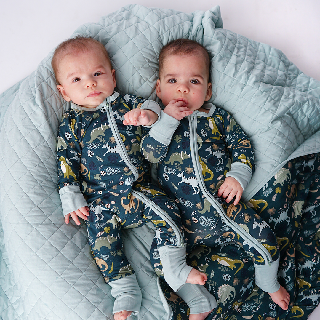 two babies lay on the "prehistoric friends" quilt. both babies are in the "prehistoric friends" convertible.the "prehistoric friends" print is a mix of cute dinosaurs, eggs, and branches, all scattered on a dark green background.