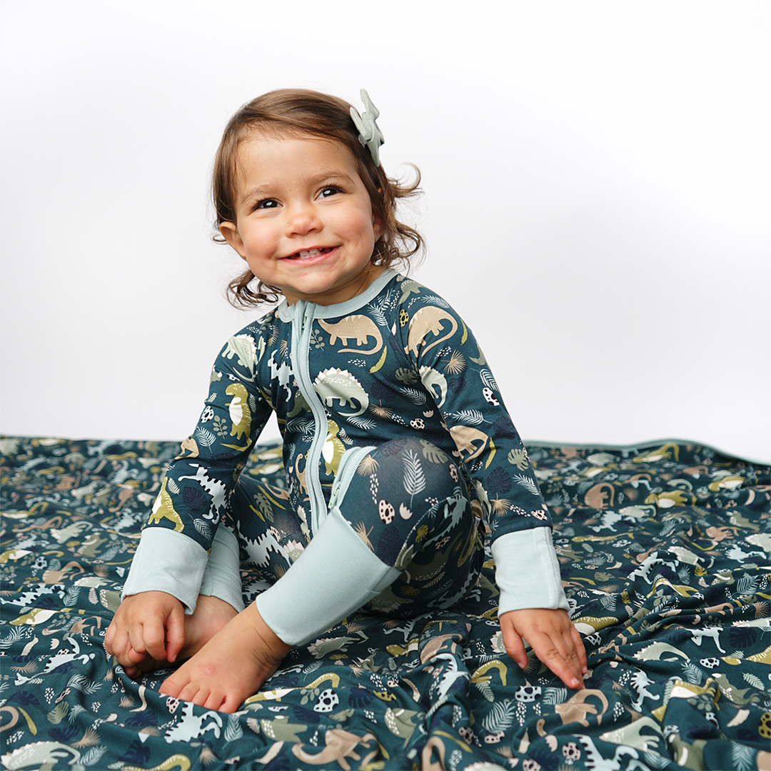 a young toddler girl sits on the "prehistoric friends" blanket. she is wearing the "prehistoric friends" convertible. the "prehistoric friends" print is a mix of cute dinosaurs, eggs, and branches, all scattered on a dark green background.