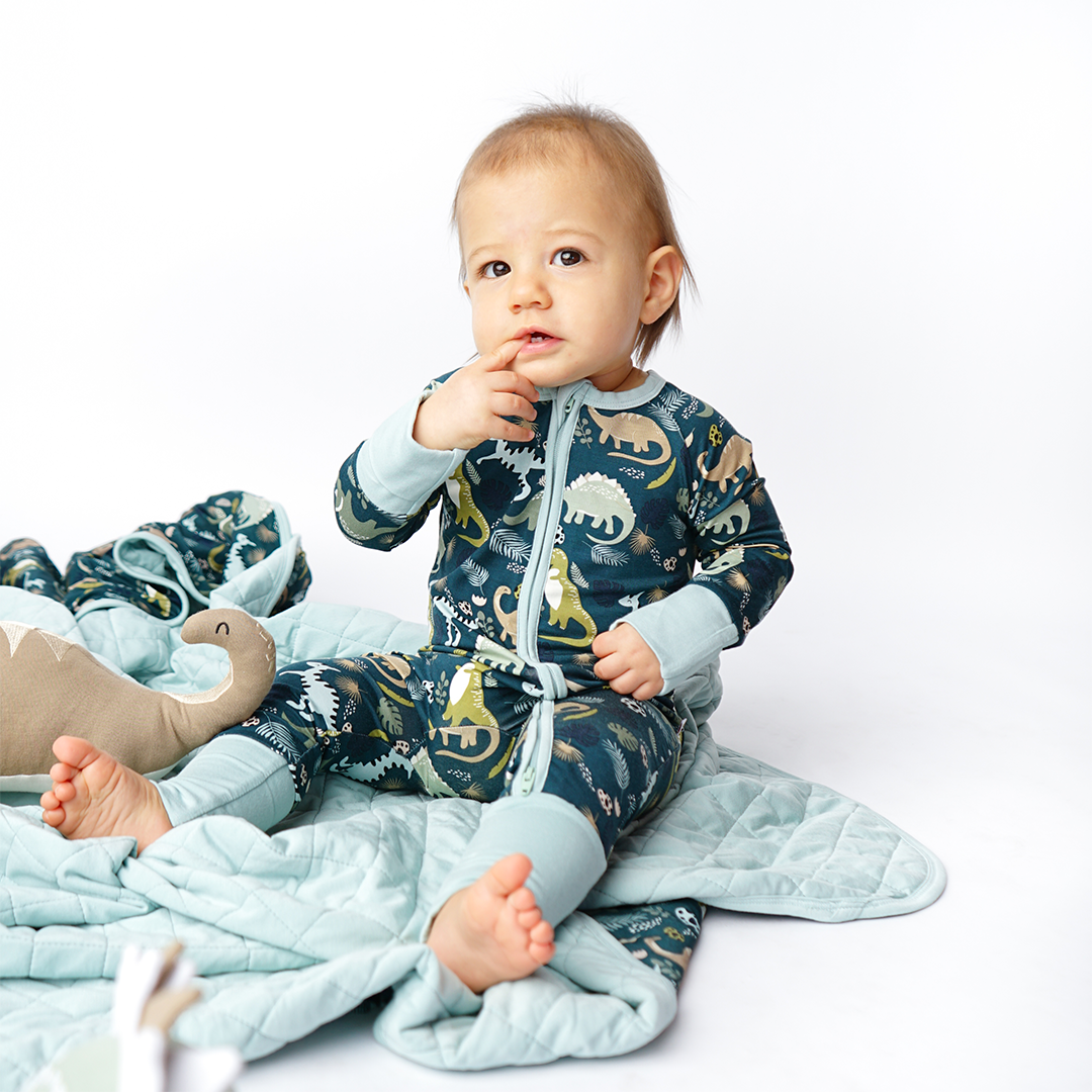 a baby sits on the "prehistoric friends" quilt with his finger in his mouth. the baby wears the "prehistoric friends" convertible. the "prehistoric friends" print is a mix of cute dinosaurs, eggs, and branches, all scattered on a dark green background.