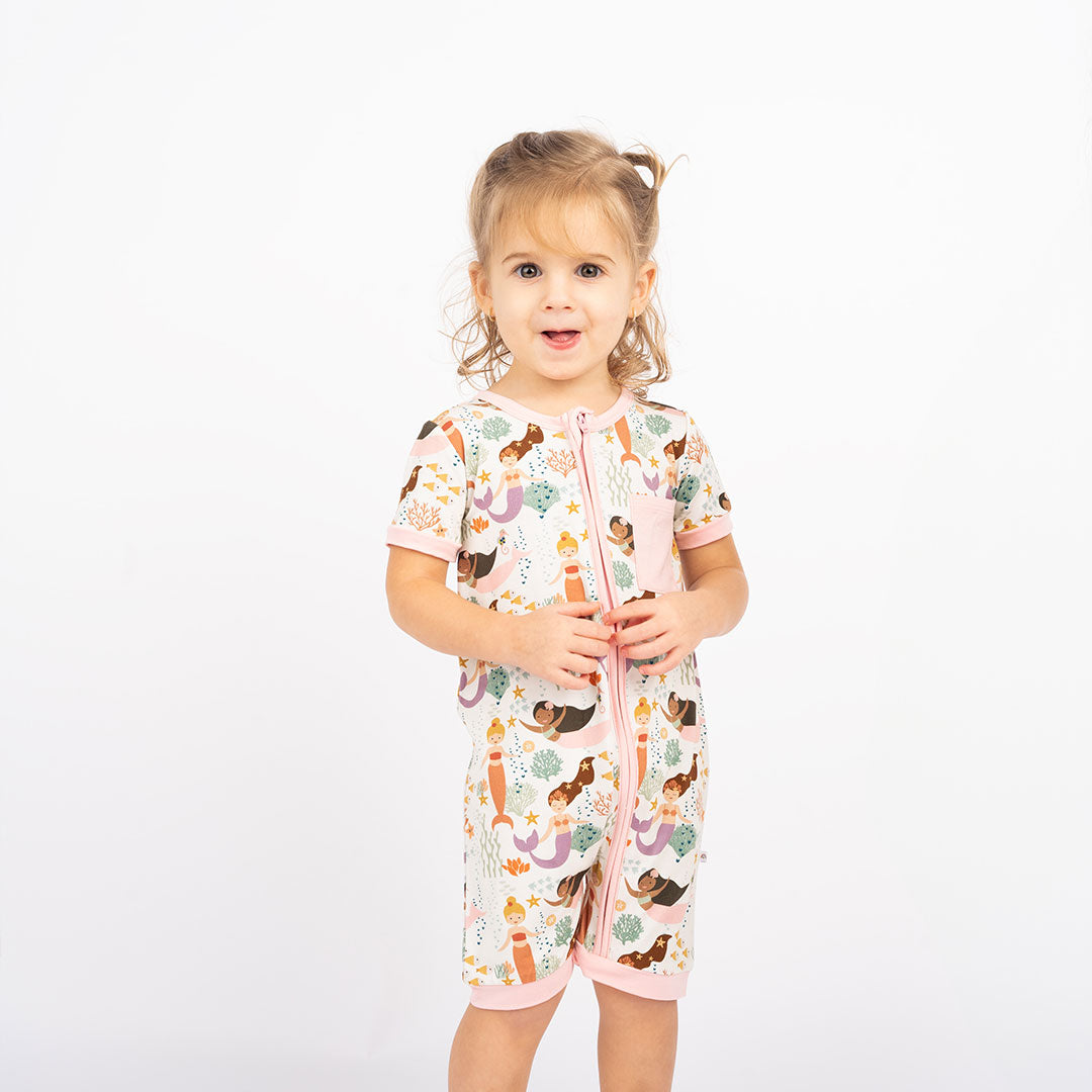 girl poses in her "making waves" shortie. the "making waves" print has a diverse spread of mermaids, sea coral, starfish, fish, and bubbles all spread out in different colors. this is all put on a beige background.