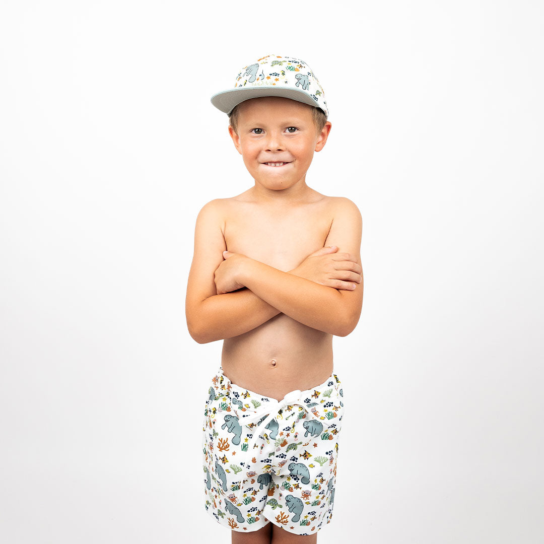 a boy stands with his arms crossed. he is wearing the "manatee" boy swim trunks and the matching snapback hat. the print "manatee" unites the florida sea life in a majestic print. there is a variation of florida manatees, yellow and pink sea horses, yellow and pink starfish, bubbles, turtles, and coral patterned across a white background