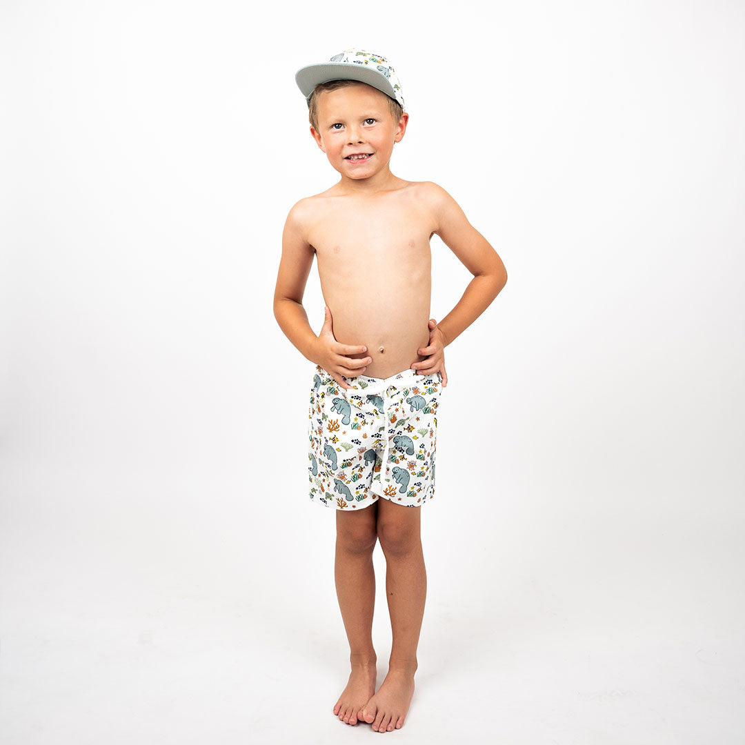 boy stands with his hands on his hips. he is wearing the "manatee" boy swim trunks and the matching snapback hat. the print "manatee" unites the florida sea life in a majestic print. there is a variation of florida manatees, yellow and pink sea horses, yellow and pink starfish, bubbles, turtles, and coral patterned across a white background
