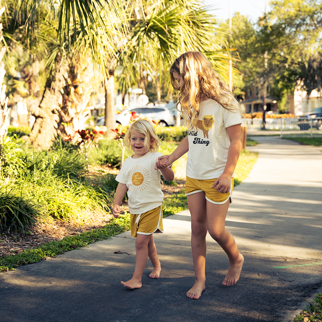 2 girls walk on a sidewalk hand in hand. they are wearing the golden sunshine bamboo terry track shorts. the younger one is wearing the "here comes the sun" tee. the older one is wearing the "wild things" tee. they are outside and you can see grass, palm trees, and a parking lot behind them.