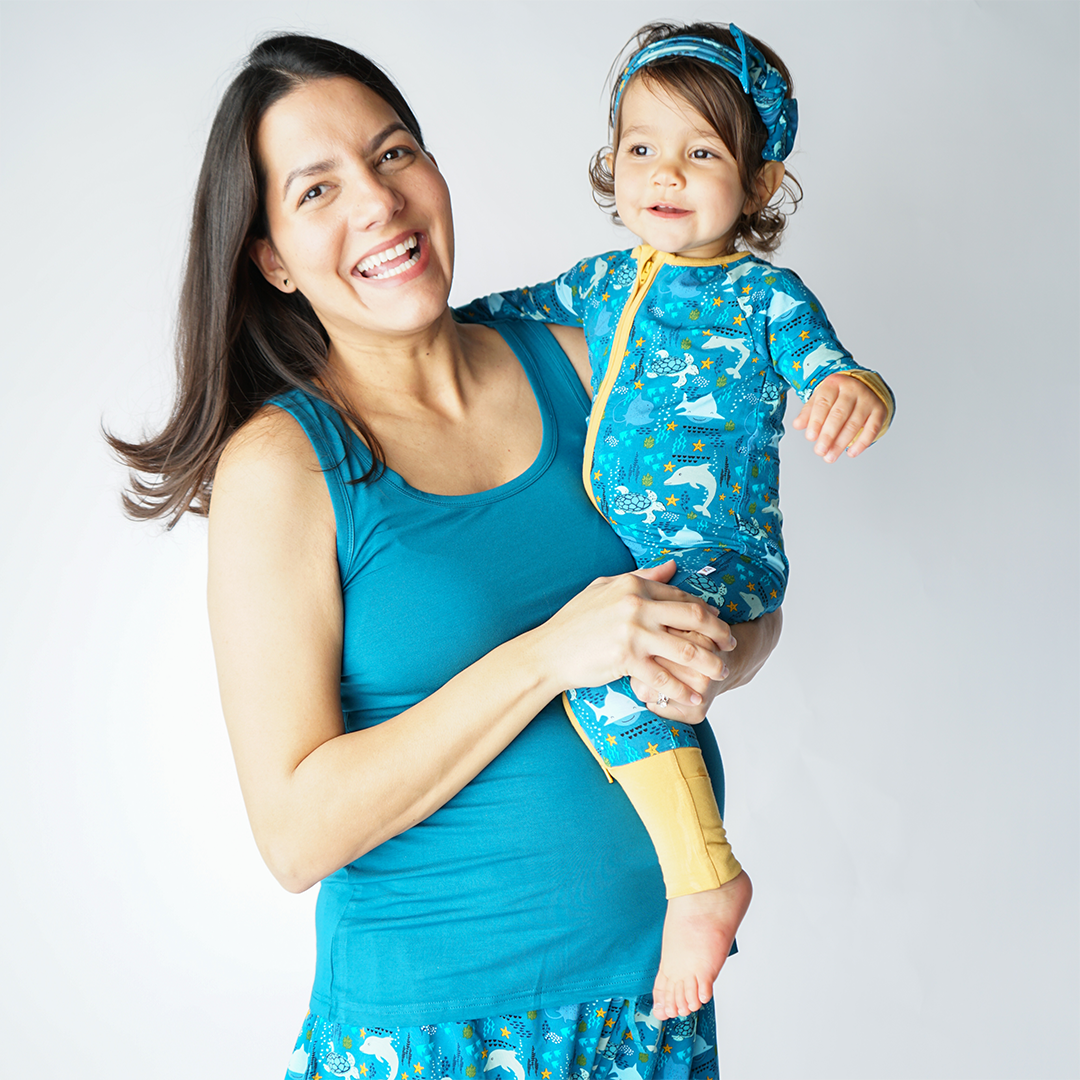 mom holds her baby on her hip. she is wearing the "ocean friends" bamboo women's shorts and the "ocean blue" tank top. the baby smiles while wearing the "ocean friends" convertible. the "ocean friends" print is a combination of dolphins, stingrays, fish, starfish, coral, bubbles, and sharks, all spread out on a deep sea blue background.