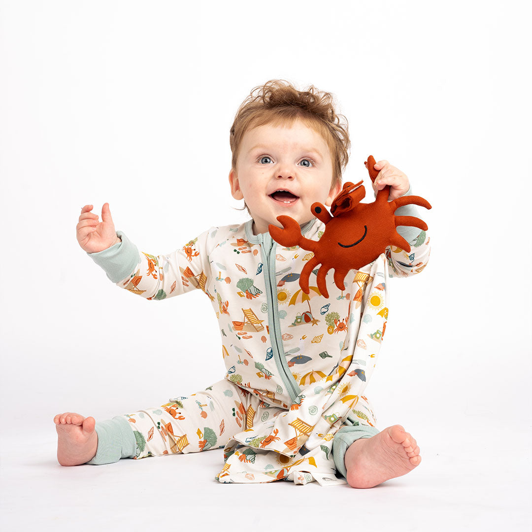 a baby sits on the floor wearing his "beach days" convertible. he is holding the "beach days" lovey. the "beach days" print is a scattered print of beach umbrellas, beach balls, sand buckets and shovels, coral, shells, beach chairs, seagulls, flipflops, and sunglass mixed in a orderly and creative way.