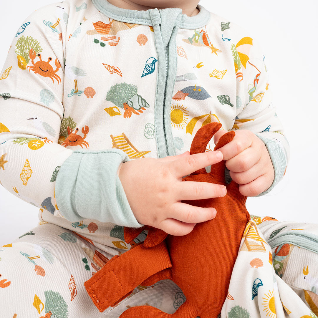 a close up picture of the "beach days" convertible and lovey. the "beach days" print is a scattered print of beach umbrellas, beach balls, sand buckets and shovels, coral, shells, beach chairs, seagulls, flipflops, and sunglass mixed in a orderly and creative way.