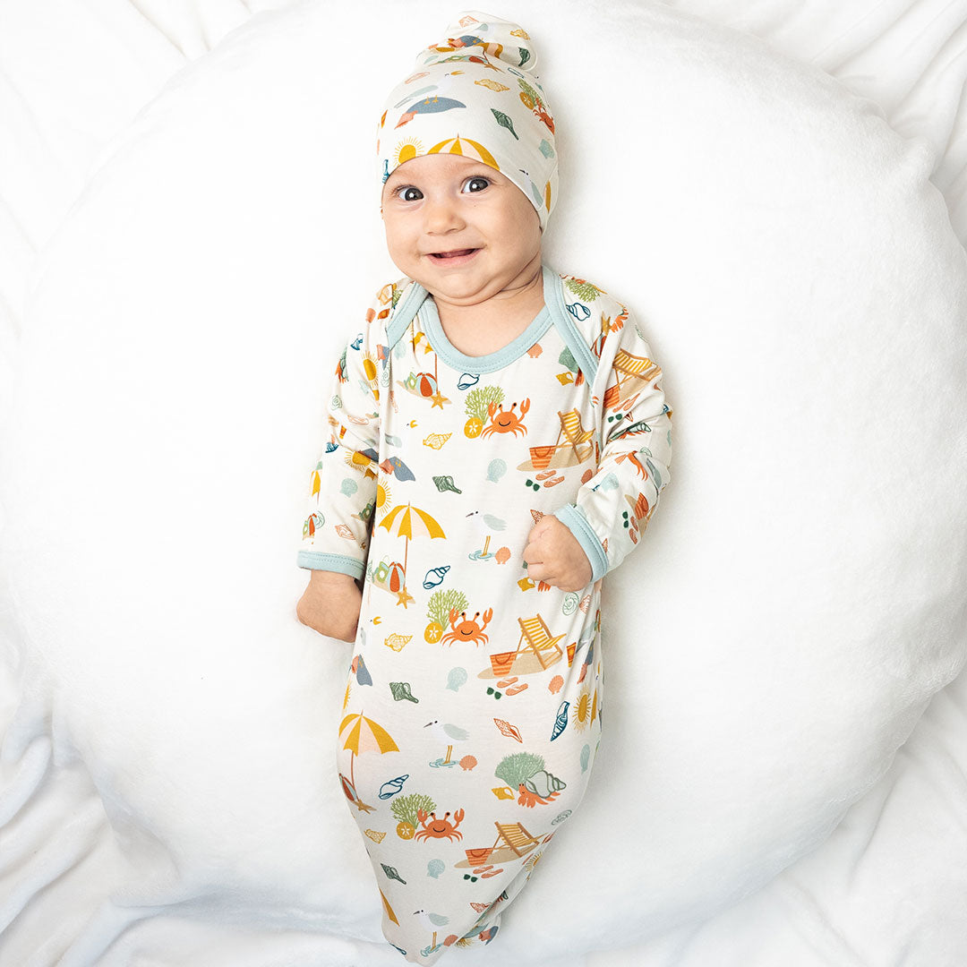 a baby smiles cheerfully in his "beach days" gown and hat set. he lays on his back on a white pillow. the "beach days" print is a scattered print of beach umbrellas, beach balls, sand buckets and shovels, coral, shells, beach chairs, seagulls, flipflops, and sunglass mixed in a orderly and creative way.