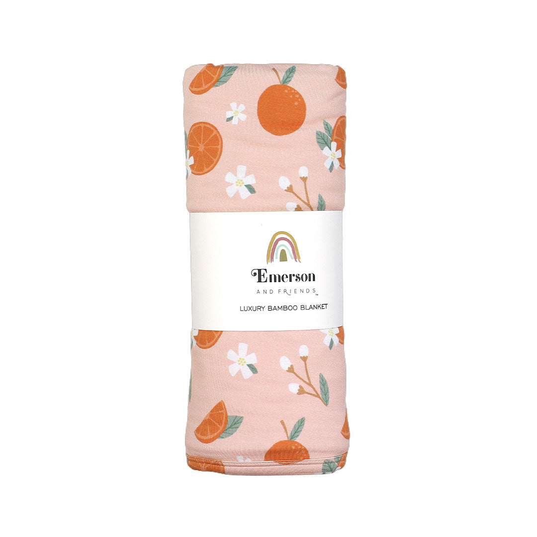 the "freshly squeezed" bamboo blanket. the "freshly squeezed" print has an assortment of full and half cut oranges scattered around. there is also flower heads and flower stems that intermingle within the print. this is all space out around a pink background space.